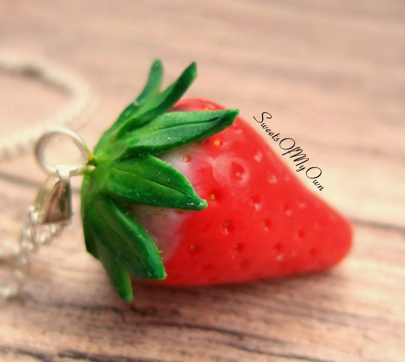 May include: A red and green polymer clay strawberry pendant on a silver chain. The strawberry has a white tip and a green leafy top. The pendant is labeled "Sweets Of My Own".