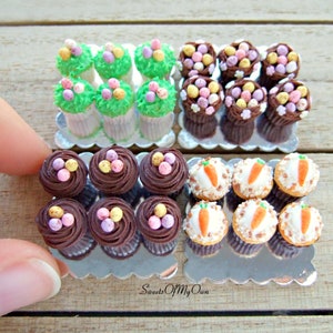 Peut inclure: Trois plateaux de mini-cupcakes d&eacute;cor&eacute;s sur le th&egrave;me de P&acirc;ques. Le premier plateau pr&eacute;sente des cupcakes avec un gla&ccedil;age vert et des &oelig;ufs en bonbons de couleur pastel. Le deuxi&egrave;me plateau pr&eacute;sente des cupcakes avec un gla&ccedil;age au chocolat et des &oelig;ufs en bonbons de couleur pastel. Le troisi&egrave;me plateau pr&eacute;sente des cupcakes avec un gla&ccedil;age blanc et des d&eacute;corations de carottes orange. Les plateaux sont sur une surface en bois. Sweets Of My Own.