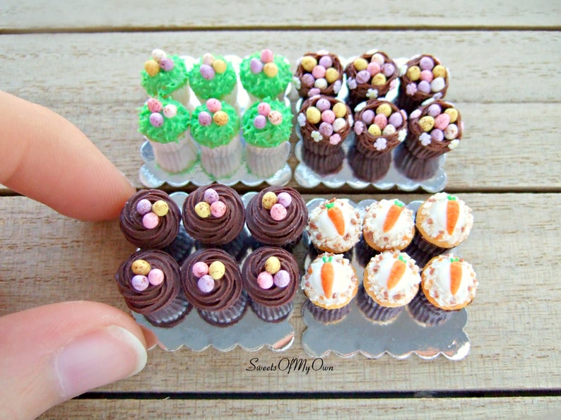 Peut inclure: Trois plateaux de mini-cupcakes d&eacute;cor&eacute;s sur le th&egrave;me de P&acirc;ques. Le premier plateau pr&eacute;sente des cupcakes avec un gla&ccedil;age vert et des &oelig;ufs en bonbons de couleur pastel. Le deuxi&egrave;me plateau pr&eacute;sente des cupcakes avec un gla&ccedil;age au chocolat et des &oelig;ufs en bonbons de couleur pastel. Le troisi&egrave;me plateau pr&eacute;sente des cupcakes avec un gla&ccedil;age blanc et des d&eacute;corations de carottes orange. Les plateaux sont sur une surface en bois. Sweets Of My Own.