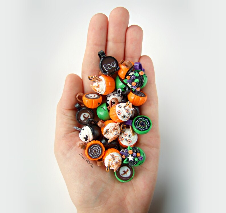 May include: A hand holding a collection of Halloween-themed charms, featuring pumpkins, ghosts, bats, and spiders. The charms are crafted from polymer clay and are painted in a range of colours, including orange, green, black, and white. Some of the charms have intricate details, such as eyes, mouths, and wings.