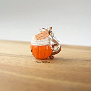 May include: A small orange pumpkin-shaped charm with white frosting and a waffle cookie on top. The charm has a silver clip attached to it.