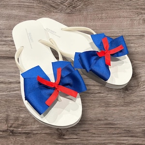 May include: White flip-flops with blue bows and red accents. The flip-flops have a simple design with a thong strap. The bows are attached to the top of the flip-flops, adding a decorative touch. The flip-flops are on a wooden surface.