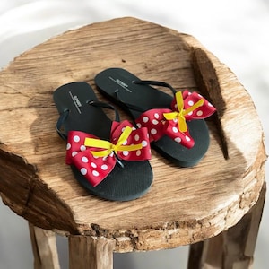 May include: A pair of dark green flip-flops with red bows featuring white polka dots and yellow accents. The flip-flops are displayed on a rustic wooden surface.