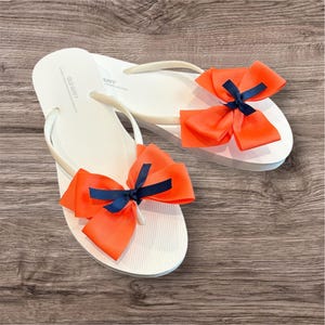May include: White flip-flops with orange bows and navy blue accents. The flip-flops have white straps and soles. The bows are attached to the top of the flip-flops. The flip-flops are on a wooden surface.