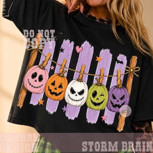 May include: A black t-shirt featuring a graphic of five colorful jack-o'-lanterns hanging from a clothesline. The pumpkins are pink, orange, white, green, and purple, each with a unique face. The design includes purple vertical brush strokes, small stars, and a heart.