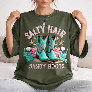 Może przedstawiać: Zielona koszulka z haftowanym napisem "SALTY HAIR SANDY BOOTS". Koszulka przedstawia turkusowe kowbojki, r&oacute;żowe kwiaty, rozgwiazdy i muszle.