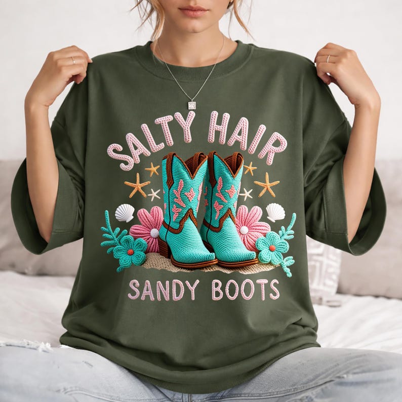 Może przedstawiać: Zielona koszulka z haftowanym napisem "SALTY HAIR SANDY BOOTS". Koszulka przedstawia turkusowe kowbojki, r&oacute;żowe kwiaty, rozgwiazdy i muszle.