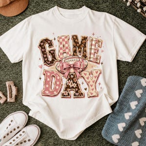 K&ouml;nnte beinhalten: Ein wei&szlig;es Kurzarm-T-Shirt mit dem Schriftzug GAME DAY in gro&szlig;en Blockbuchstaben. Die Buchstaben enthalten Muster aus braunen Footballs, rosa Karos und Blumen. Eine zentrale Football-Grafik ist mit einer gro&szlig;en rosa Schleife verziert.