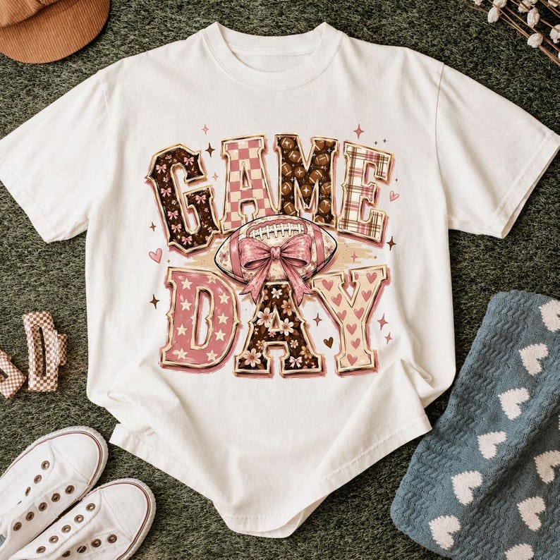 K&ouml;nnte beinhalten: Ein wei&szlig;es Kurzarm-T-Shirt mit dem Schriftzug GAME DAY in gro&szlig;en Blockbuchstaben. Die Buchstaben enthalten Muster aus braunen Footballs, rosa Karos und Blumen. Eine zentrale Football-Grafik ist mit einer gro&szlig;en rosa Schleife verziert.