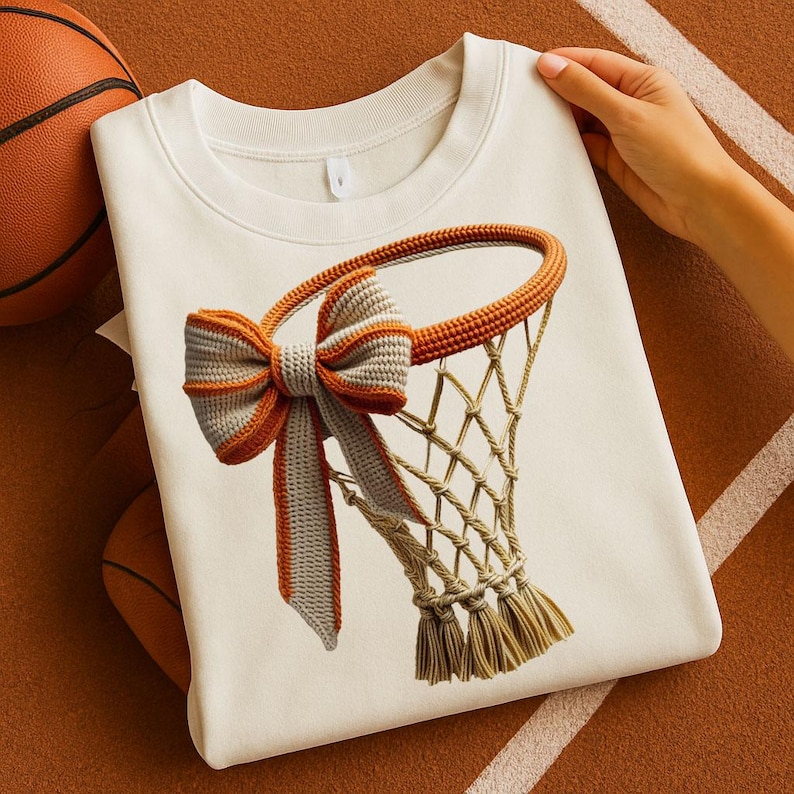 May include: Cream-colored sweatshirt featuring a basketball net design with a bow. The net and bow are detailed with orange and gray accents. A basketball is visible in the background. The sweatshirt is made of a soft material.