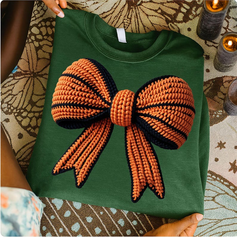 May include: A green sweatshirt featuring a large, crocheted bow design. The bow is primarily orange with black stripes and detailing. The sweatshirt is a solid green color, and the bow is the focal point of the design.