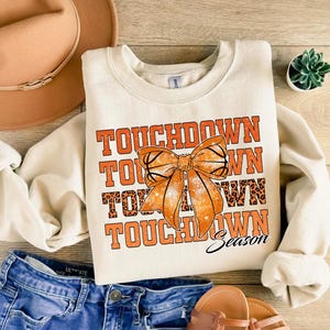 Może przedstawiać: Kremowa bluza z napisem "TOUCHDOWN" w pomarańczowych literach, grafiką kokardy i słowem "Season". Kokarda ma wz&oacute;r koszyk&oacute;wki. Bluza jest połączona z niebieskimi szortami dżinsowymi, brązowym kapeluszem i sandałami.