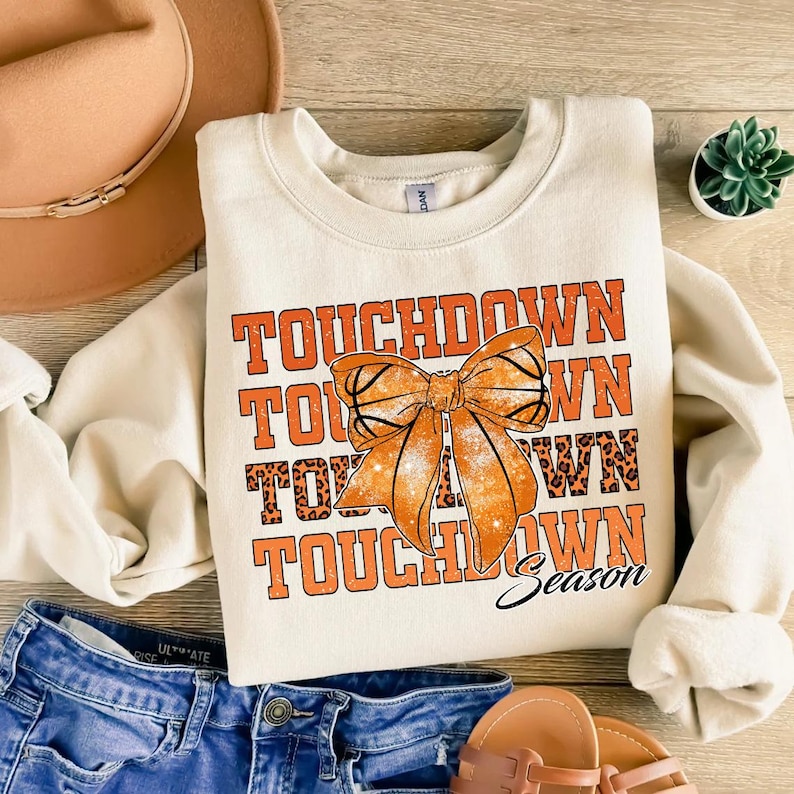 Może przedstawiać: Kremowa bluza z napisem "TOUCHDOWN" w pomarańczowych literach, grafiką kokardy i słowem "Season". Kokarda ma wz&oacute;r koszyk&oacute;wki. Bluza jest połączona z niebieskimi szortami dżinsowymi, brązowym kapeluszem i sandałami.