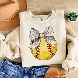 May include: A cream-colored sweatshirt with a graphic of a yellow softball with a white and gold bow on top.