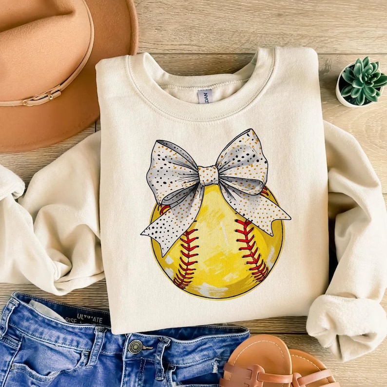 May include: A cream-colored sweatshirt with a graphic of a yellow softball with a white and gold bow on top.