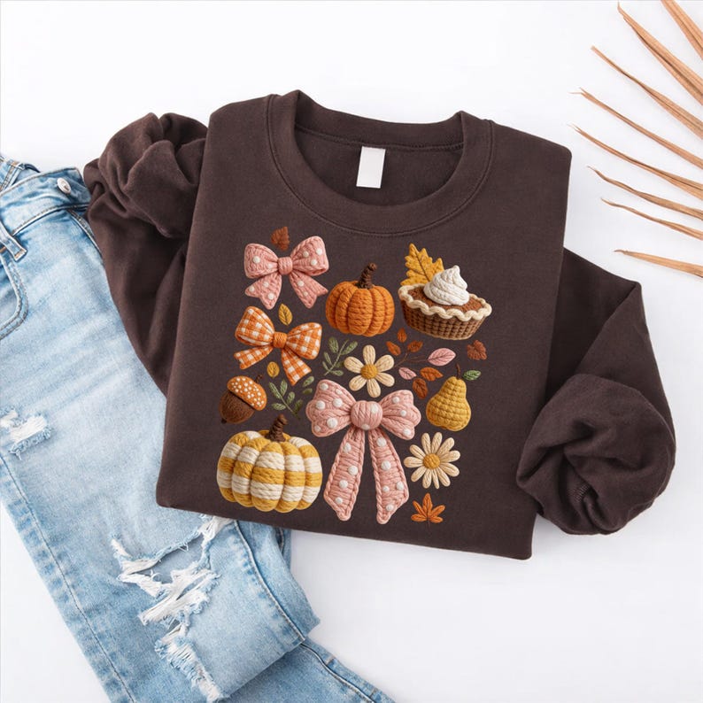 May include: A brown sweatshirt featuring an embroidered fall-themed design. The design includes pumpkins, bows, flowers, and a pie. The sweatshirt is paired with light blue jeans.