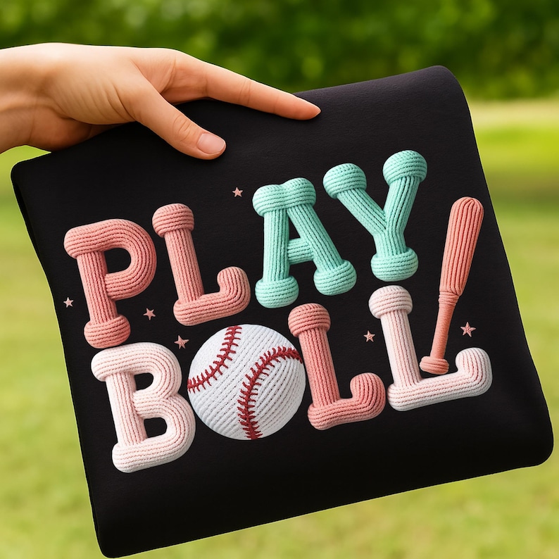 May include: Black pillow with the words "PLAY BALL" in a 3D knitted style. The letters are pink and teal, with a baseball and a baseball bat. The baseball is white with red stitching. Small star accents are scattered around the text.