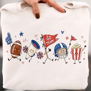 K&ouml;nnte beinhalten: Ein cremefarbenes Sweatshirt mit einem Grafikdruck von animierten Sportartikeln, darunter ein Football mit einem Nummer-eins-Schaumstofffinger, ein Megafon, Pompons, eine rote Flagge mit dem Text GO TEAM!, ein Footballhelm und Popcorn.