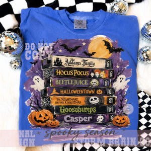 May include: A blue crewneck t-shirt featuring a graphic print of a stack of seven VHS tapes labeled with movie titles including The Addams Family, Hocus Pocus, Beetlejuice, Halloweentown, The Nightmare Before Christmas, Goosebumps, and Casper.