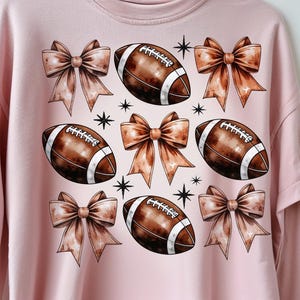 May include: Pink sweatshirt featuring a repeating pattern of brown footballs, bows, and starbursts. The footballs have white stripes and laces. The bows are a light brown color. The starbursts are black.