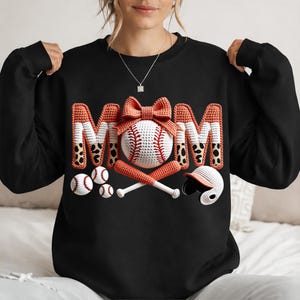 May include: Black sweatshirt with a baseball-themed design. The word "MOM" is spelled out in orange and white letters with a leopard print pattern. A baseball, bats, and a helmet are also featured.