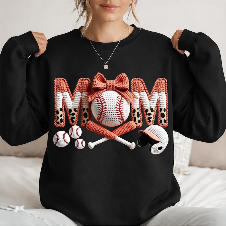 May include: Black sweatshirt with a baseball-themed design. The word "MOM" is spelled out in orange and white letters with a leopard print pattern. A baseball, bats, and a helmet are also featured.