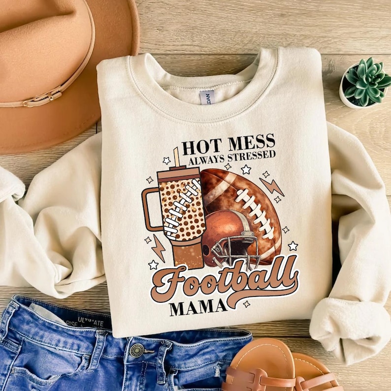 May include: Cream-colored sweatshirt with a football-themed graphic. The design includes a football, helmet, and a tumbler with the text "HOT MESS ALWAYS STRESSED Football MAMA". The sweatshirt is paired with blue jeans and a brown hat.