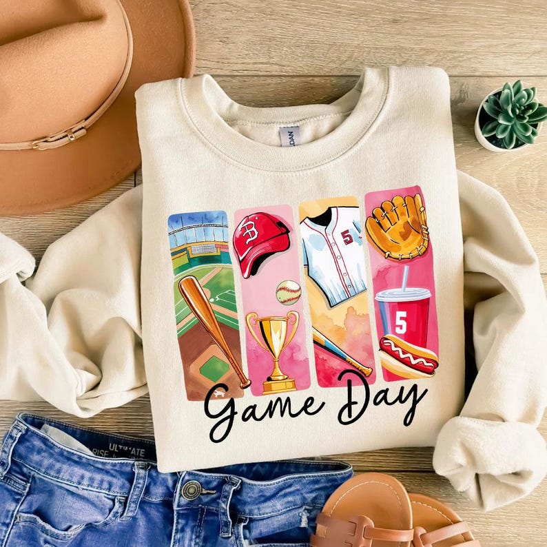 May include: A beige sweatshirt with a graphic print of baseball-themed items, including a baseball bat, a baseball glove, a baseball cap, a trophy, a hot dog, and a drink. The text "Game Day" is printed below the graphic.