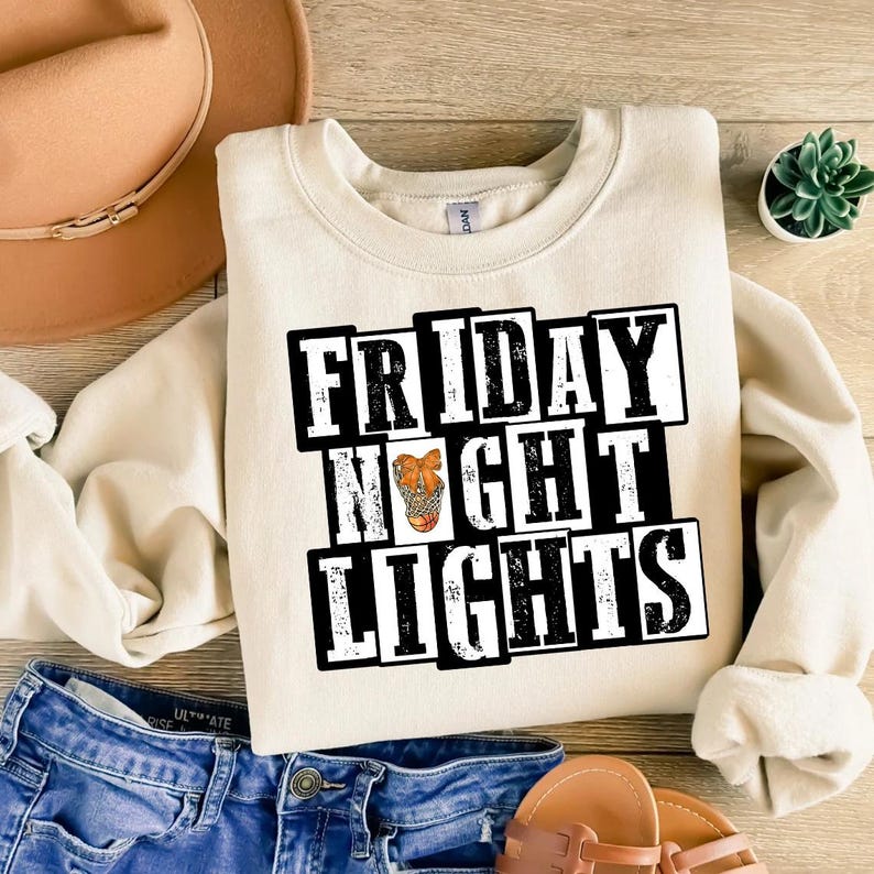 Może przedstawiać: Kremowa bluza z napisem "FRIDAY NIGHT LIGHTS" w czarno-białych literach blokowych. Litera "O" w słowie "NIGHT" przedstawia koszyk&oacute;wkę w siatce. Bluza jest połączona z niebieskimi szortami dżinsowymi i brązowym kapeluszem.