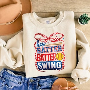 May include: A cream-colored sweatshirt with a red, white, and blue graphic design that says "Hey Batter Batter Swing" with a baseball bat and a softball.
