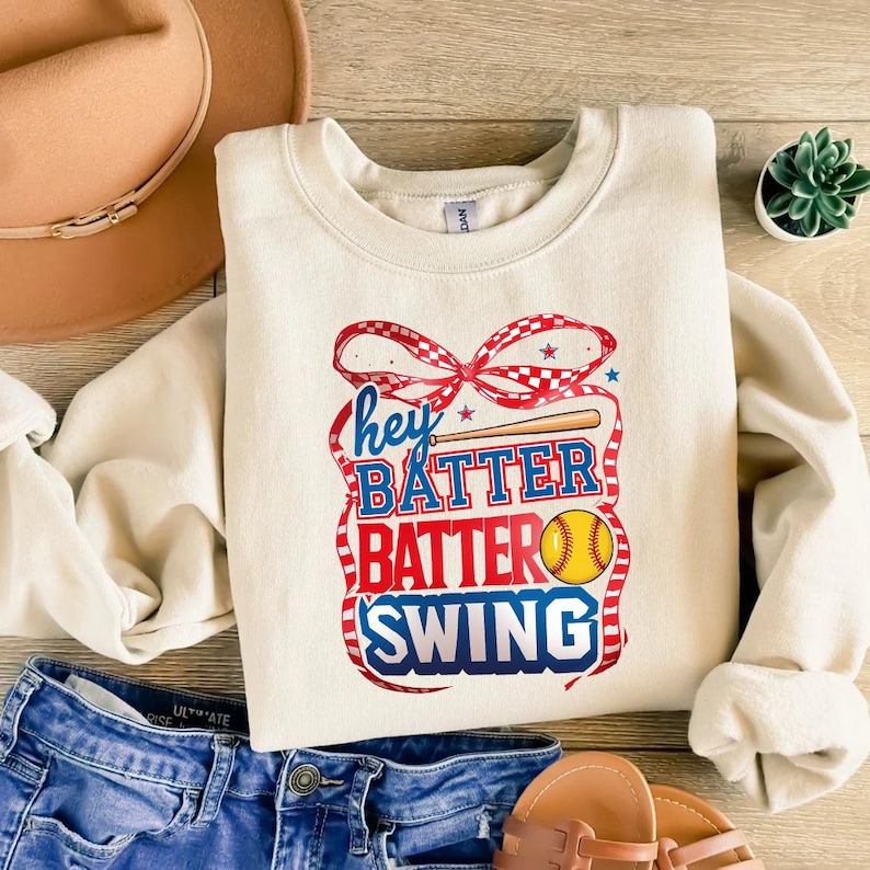 May include: A cream-colored sweatshirt with a red, white, and blue graphic design that says "Hey Batter Batter Swing" with a baseball bat and a softball.
