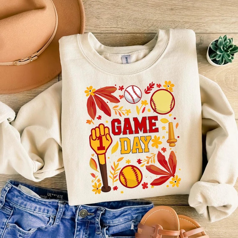 May include: A cream colored sweatshirt with a graphic design of a baseball bat, baseballs, and leaves. The text "Game Day" is printed in red and yellow letters.