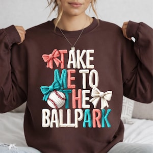 May include: A brown sweatshirt with the phrase "TAKE ME TO THE BALLPARK" in white, coral, teal, and cream-colored 3D lettering. A baseball and bows in matching colors are also featured in the design.