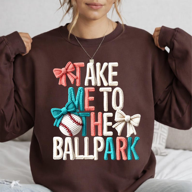 May include: A brown sweatshirt with the phrase "TAKE ME TO THE BALLPARK" in white, coral, teal, and cream-colored 3D lettering. A baseball and bows in matching colors are also featured in the design.