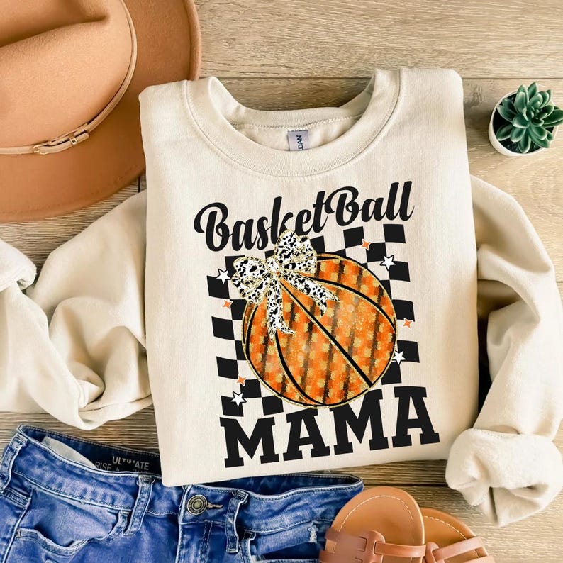 Może przedstawiać: Kremowa bluza z napisem "BasketBall MAMA" w czarnym skrypcie i blokowych literach. Grafika koszyk&oacute;wki z kokardą i szachownicą jest wyśrodkowana z przodu. Bluza jest połączona z niebieskimi dżinsowymi szortami i brązowym kapeluszem.