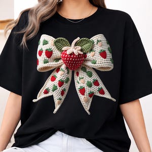 Może przedstawiać: Czarny t-shirt z dużym, kremowym, szydełkowym łukiem ozdobionym czerwonymi truskawkami i zielonymi liśćmi. Duża, czerwona szydełkowa truskawka znajduje się na środku łuku. Koszulka ma kr&oacute;tkie rękawy i okrągły dekolt.