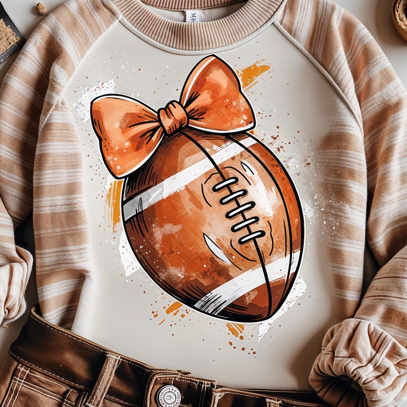 May include: A beige sweatshirt with a watercolor-style football graphic and an orange bow. The football is brown with white stripes and laces. The sweatshirt has tan and white striped sleeves and is paired with brown pants.