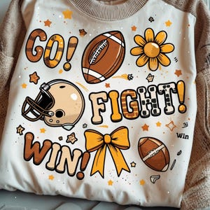 May include: Cream-colored sweatshirt with a football theme. The design includes the words "GO! FIGHT! WIN!" in a playful font, along with footballs, a helmet, a bow, and star and flower accents. The sweatshirt has a beige collar and sleeves.