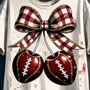 May include: White t-shirt featuring a whimsical design of two cherries styled as footballs, complete with white laces and a gold glitter outline. A large, plaid bow with gold trim sits above the cherries, creating a playful and unique graphic.