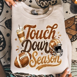 K&ouml;nnte beinhalten: Ein beigefarbenes Sweatshirt mit Rundhalsausschnitt und einem braunen Aufdruck mit dem Text "Touch Down Season". Das Design zeigt einen Football, einen Becher mit Strohhalm und einen Footballhelm mit einem Herzsymbol an der Seite in Braunt&ouml;nen.