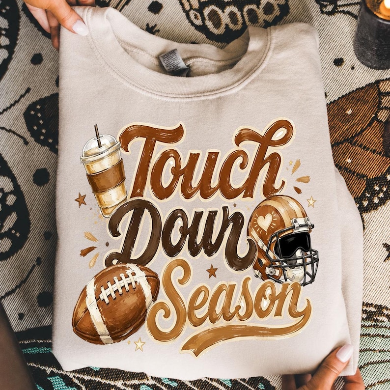 K&ouml;nnte beinhalten: Ein beigefarbenes Sweatshirt mit Rundhalsausschnitt und einem braunen Aufdruck mit dem Text "Touch Down Season". Das Design zeigt einen Football, einen Becher mit Strohhalm und einen Footballhelm mit einem Herzsymbol an der Seite in Braunt&ouml;nen.