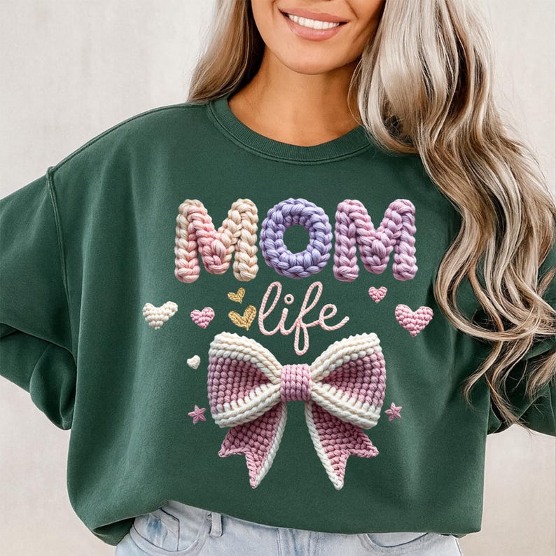 May include: Dark green sweatshirt with the words "MOM life" in a crochet style. The "MOM" is in pink and purple, and the word "life" is in cursive. A large crochet bow and hearts decorate the front.