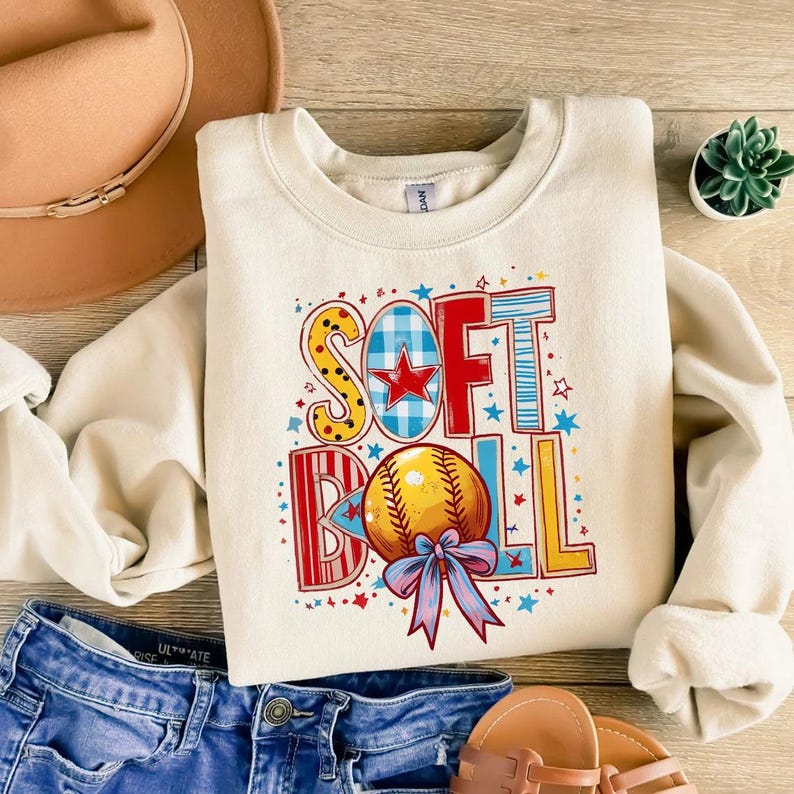 May include: A cream-colored sweatshirt with a colorful graphic design that says "SOFTBALL" with a red star and a yellow and white baseball.