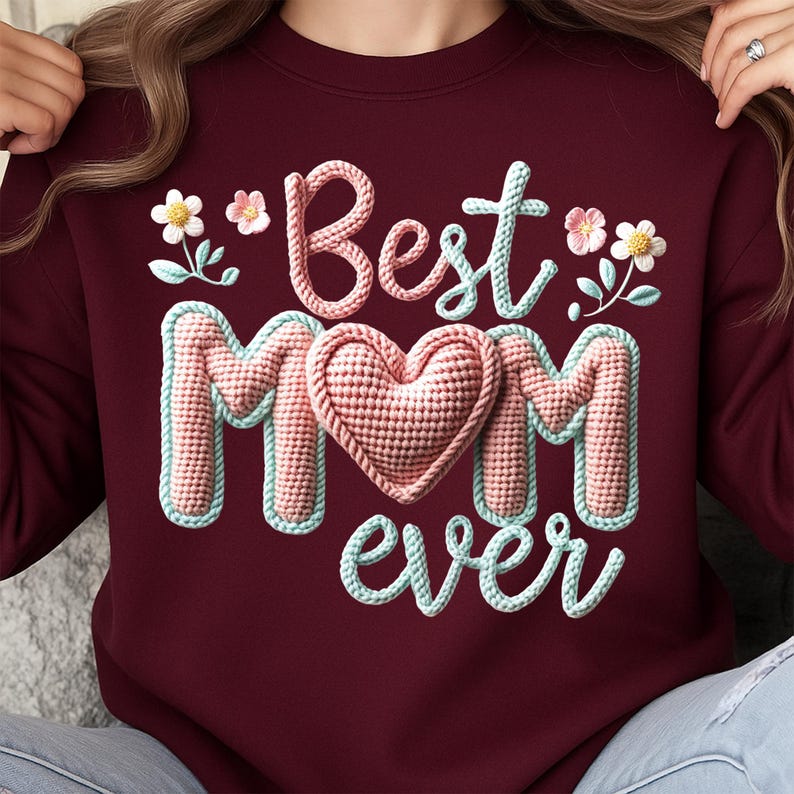May include: Burgundy sweatshirt with the words "Best Mom Ever" in a 3D textured design. The text is in white, pink, and light blue, with a pink heart in the center of the word "Mom". Floral accents are on the top left and right.