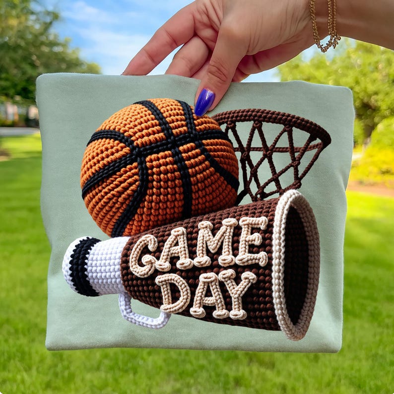 May include: A light green t-shirt featuring a crochet basketball, hoop, and megaphone. The basketball is brown and orange with black stripes. The megaphone is brown with the words "GAME DAY" in cream-colored letters.