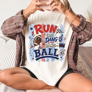 K&ouml;nnte beinhalten: Ein cremefarbenes T-Shirt mit einem Grafikdruck und dem Text RUN THE DANG DANG BALL. Das Design zeigt einen Cartoon-Football mit Sonnenbrille und Turnschuhen, eine Zielflagge, einen blauen Schaumstofffinger, eine Anzeigetafel und eine silberne Pfeife.