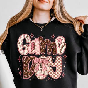 K&ouml;nnte beinhalten: Ein schwarzes Sweatshirt mit Rundhalsausschnitt und dem Schriftzug Game Day in einer stilisierten Schriftart. Die Buchstaben sind mit rosa und braunen Mustern verziert, darunter Fu&szlig;b&auml;lle, Schleifen, Blitze und Karos, akzentuiert durch kleine rosa Sterne.