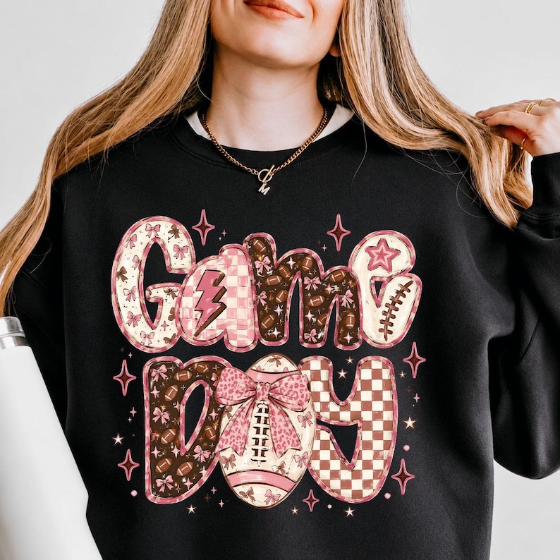 K&ouml;nnte beinhalten: Ein schwarzes Sweatshirt mit Rundhalsausschnitt und dem Schriftzug Game Day in einer stilisierten Schriftart. Die Buchstaben sind mit rosa und braunen Mustern verziert, darunter Fu&szlig;b&auml;lle, Schleifen, Blitze und Karos, akzentuiert durch kleine rosa Sterne.