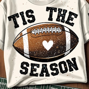 May include: Cream-colored t-shirt with a football graphic. The football is brown with white stripes and a white heart. The text "TIS THE SEASON" is in black, arched above and below the football.