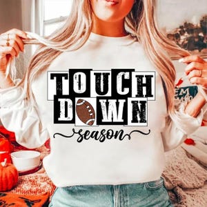 May include: White sweatshirt with the words "TOUCH DOWN" in a black and white block design, with a brown football in the center. The word "season" is written in a cursive font below the text.